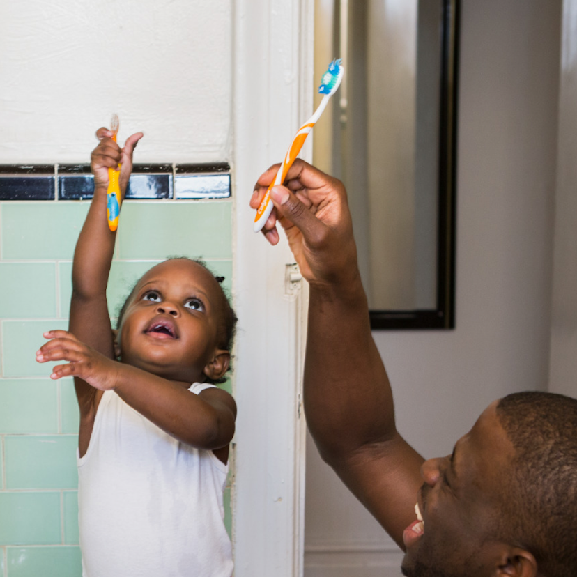 Father and child playfully lift their toothbrushes above their heads.