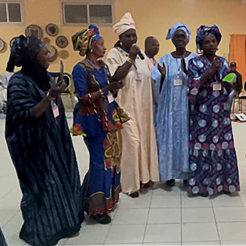 "Five African women singing at a gathering. One woman holds a green Amplio Talking Book."
